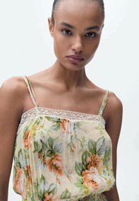Floral camisole in light beige with green and orange flowers. Features thin straps and lace trim around the neckline. Soft, textured fabric.