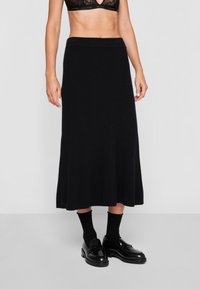 Fustă midi tricotată ribbată neagră cu margine evazată, având un brâu elastic lat. Asortată cu loafers negri și șosete.