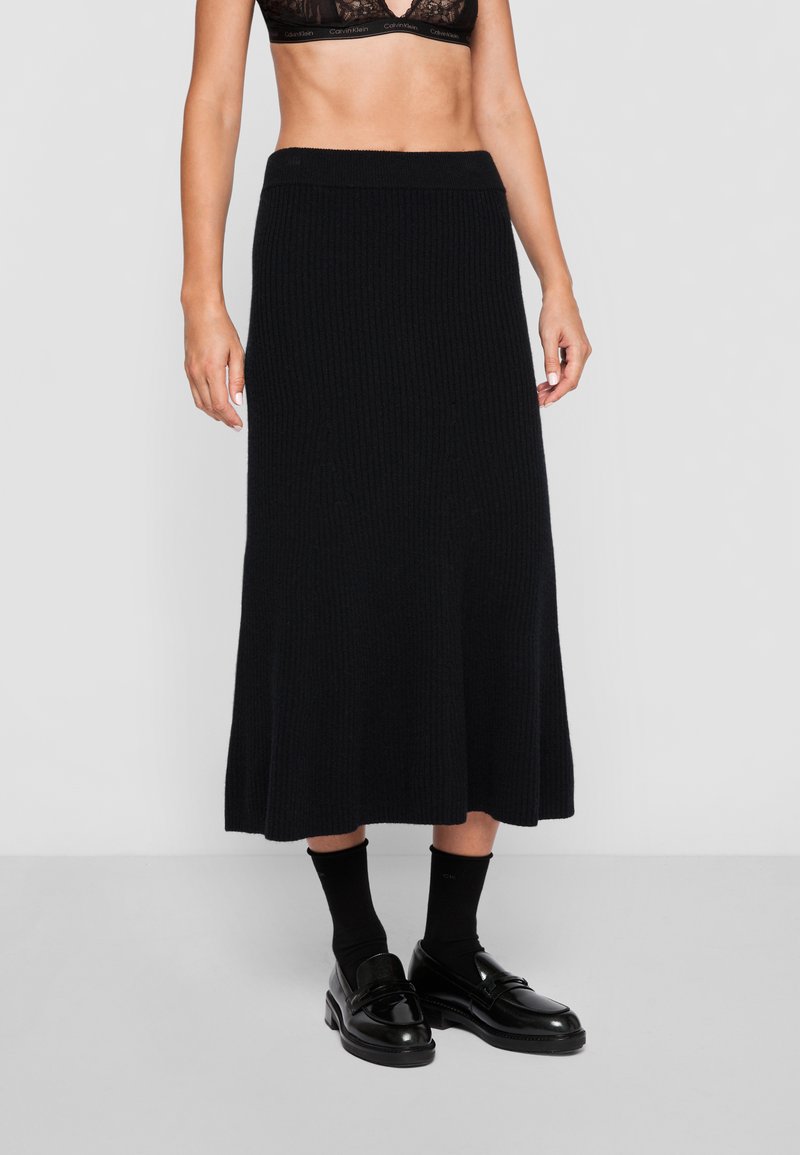 Fustă midi tricotată ribbată neagră cu margine evazată, având un brâu elastic lat. Asortată cu loafers negri și șosete.