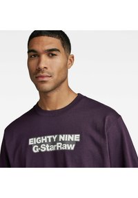 Lilla bomulls-t-skjorte med rund hals og korte ermer, med et stort grafisk trykk med teksten "EIGHTY NINE G-StarRaw" over brystet.