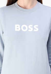 Gaiši zils džemperis ar apaļu kakla izgriezumu, uz kura priekšpuses ir lieli balti burti ar vārdu "BOSS". Mīksta auduma ar gludu tekstūru.