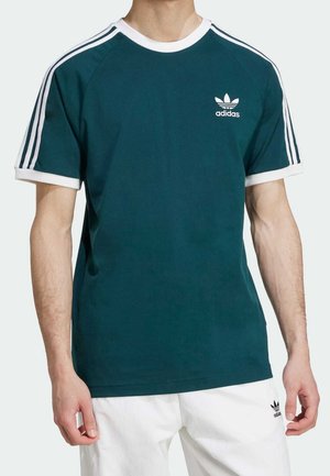 Mann trägt ein türkisfarbenes Adidas T-Shirt mit weißen Streifen auf den Schultern und Logo auf der Brust, kombiniert mit weißen Shorts mit kleinem Adidas-Logo.