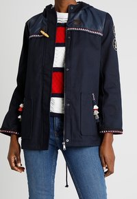 Person med en marinblå jacka med dekorativa tofsar, snodd och dragkedja, över en röd, vit och marinblå randig tröja och blå jeans.