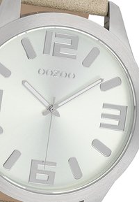 Montre argentée ronde avec de grands chiffres aux positions 3, 6, 9 et 12. Cadran métallique clair, aiguilles argentées et bracelet en cuir beige.