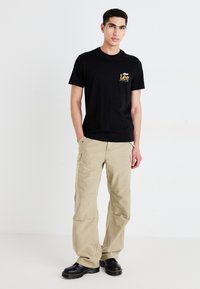 T-shirt en coton noir avec un logo "Lee" jaune sur la poitrine, associé à un pantalon cargo beige. Des bottes noires robustes montantes complètent la tenue.