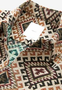 Camicia con colletto avente un motivo geometrico azteco nei toni terrosi di marrone, beige, rosso e turchese. Realizzata in tessuto testurizzato, presenta un'etichetta.