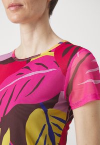 Nainen, jolla on päällään värikäs lyhythihainen paita, jossa on rohkeita vaaleanpunaisia, keltaisia, punaisia ja ruskeita abstrakteja lehtikuvioita.