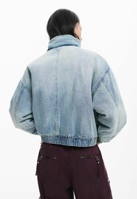 Jachetă din denim albastru deschis, cu guler înalt, siluetă supradimensionată și tiv elastic adunat. Prezintă o textură subtilă și cusături contrastante.