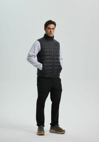 Man in een zwarte puffer vest over een lichtgrijs sweatshirt, zwarte cargo broek en bruine wandelschoenen, staand met zijn handen in zijn zakken.