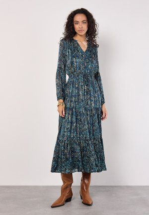 Robe maxi bleu foncé avec motif paisley, manches longues, taille froncée, jupe à volants, fabriquée en tissu léger. Bottes à talons marron.