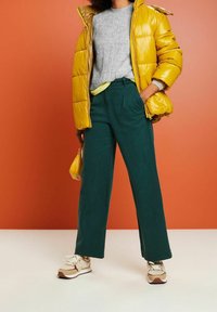 Geel gewatteerde jas, grijze gebreide trui, groene wijde broek, beige en bruine sneakers, houdend een kleine gele tas tegen een oranje achtergrond.