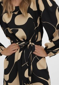 Černé a béžové šaty s vzorem, dlouhými nafouknutými rukávy a detailem na tkaničku v pase, vyrobené z hladkého materiálu a s organickými abstraktními tvary.