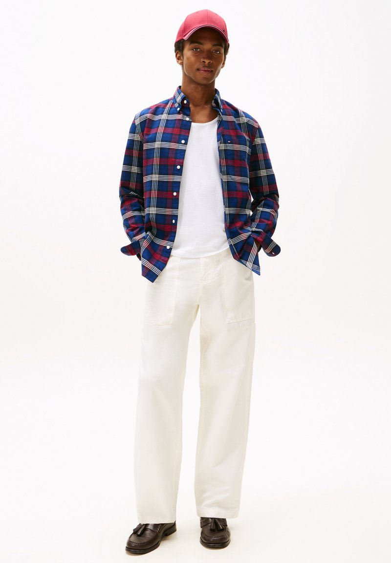 Rutigt skärp i blått, rött och vitt, bärs över en vit t-shirt. Ljust beiga vida byxor och bruna skor fulländar outfiten. Röd keps på huvudet.