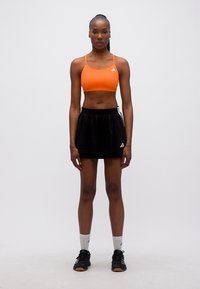 Γυναίκα που στέκεται αντιμετωπίζοντας μπροστά φορώντας πορτοκαλί αθλητικό σουτιέν, μαύρη αθλητική φούστα, λευκές κάλτσες και μαύρα αθλητικά παπούτσια, μπροστά σε λευκό φόντο.
