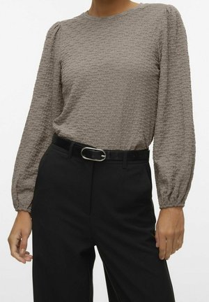 Vrouw draagt een getextureerde taupe lange mouwen top ingestopt in een zwarte hoge taille broek met een zwarte riem en zilveren gesp.
