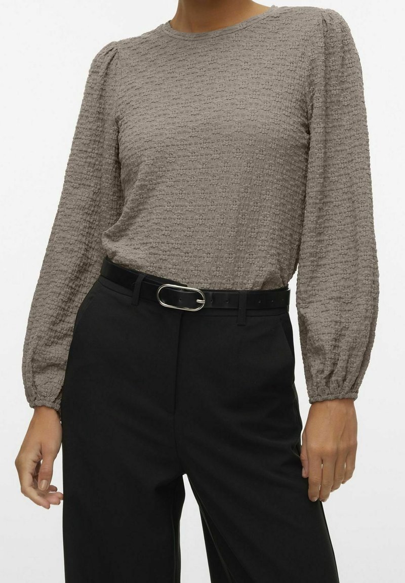 Femme portant un haut à manches longues texturé couleur taupe rentré dans un pantalon noir taille haute avec une ceinture noire et une boucle argentée.