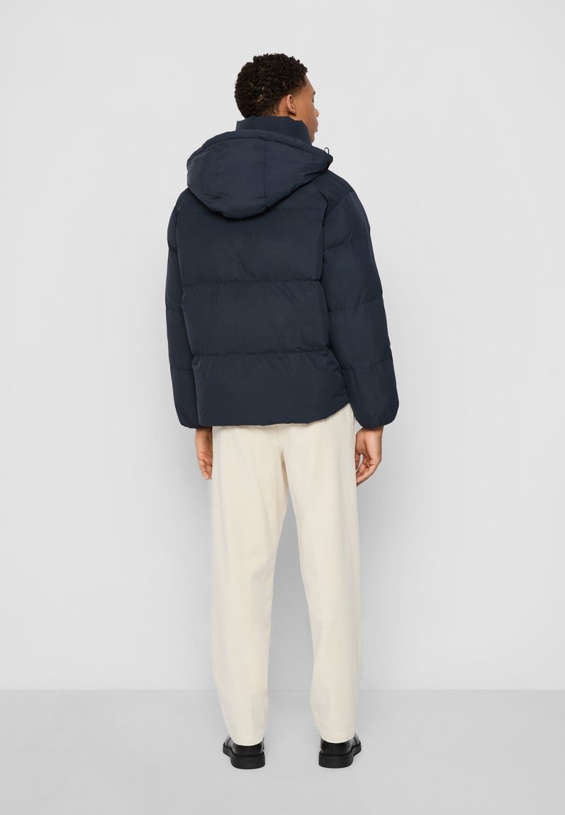 Veste matelassée navy avec capuche, au design cousu et à la coupe ample. Associée à un pantalon couleur crème et des chaussures noires.