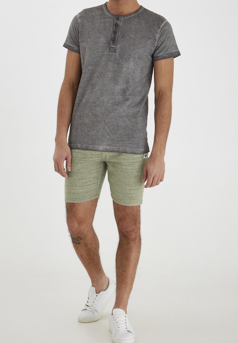 Maglietta henley grigia a maniche corte con tre bottoni marroni, abbinata a pantaloni corti verde texturizzati. Sneakers bianche completano l'outfit.