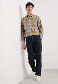 Camisa de cuadros en azul, amarillo y blanco; pantalones oscuros; cinturón marrón; zapatillas blancas. Modelo de pie contra una pared neutral.