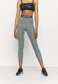Nike Pro bralette mustassa valkoisella logolla, yhdistettynä vihreisiin venyvyyden leggingsseihin, joissa on korkea vyötärö, sileä pinta ja tiukka muotoilu.