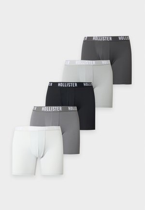 Jeune homme debout pieds nus portant un boxer noir Hollister, une main sur la hanche, sur un fond clair uni.