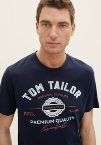 Tamno plavi pamučni T-shirt s kratkim rukavima, s bijelim i crvenim grafičkim natpisom "TOM TAILOR" i kružnim logom na prednjoj strani.