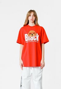 Camiseta roja de gran tamaño con el texto blanco "Black", que presenta un gráfico de un perro difuso. Mangas cortas, material de algodón y ajuste relajado.