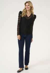 Top in pizzo nero con maniche lunghe e testurizzate, abbinato a jeans a gamba dritta blu scuro e scarpe nere basse. Design a scollo a V con un rifinitura decorativa.
