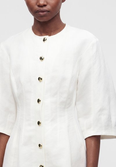 Femme portant une robe blanche avec des manches bouffantes aux demi-manches et une rangée de boutons dorés brillants sur le devant, sur un fond uni.