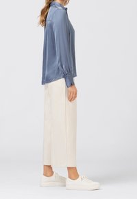 Kvinna iklädd en ljusblå satinblus, vida krämfärgade byxor och vita sneakers, stående i profil mot en enfärgad bakgrund.