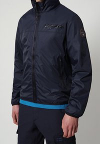 Giacca impermeabile della marina con zip frontale, colletto alto e dettaglio del logo. Presenta tasche laterali con zip e polsini elastici. Materiale leggero.