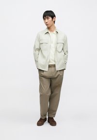 Világos bézs zipzáras dzseki krémszínű polóval, laza világosbarna nadrággal és barna tornacipővel, modell viseli, egyszerű fehér háttér előtt.