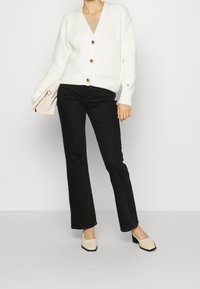 Bílý pletený kardigan s výstřihem do V a třemi knoflíky, kombinovaný s černými džíny. Modelka drží béžovou tašku a má na sobě krémové boty na blokovém podpatku.