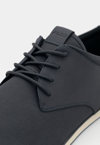 Mørkegrå herresko med snøring, teksturert overflate, glatte detaljer, gummisåle og merketag. Har flate lisser og strukturert design.