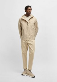 Giacca impermeabile beige con cappuccio, zip frontale e tasche laterali, abbinata a pantaloni tapered beige e sneakers beige con accenti neri.