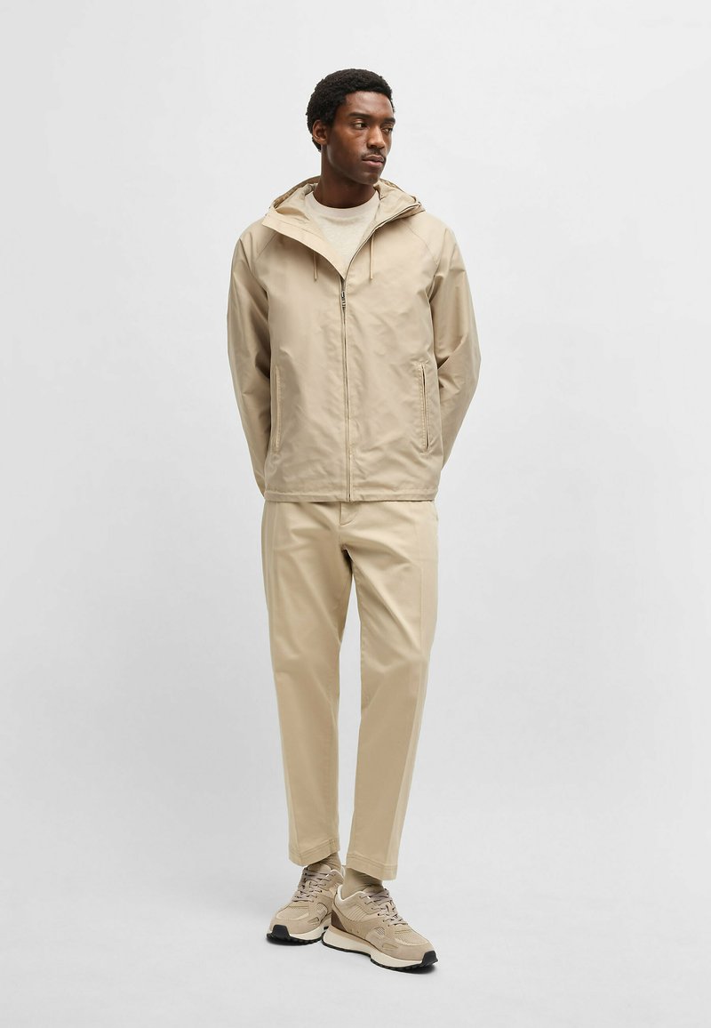 Giacca impermeabile beige con cappuccio, zip frontale e tasche laterali, abbinata a pantaloni tapered beige e sneakers beige con accenti neri.