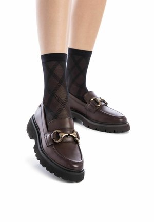 Mocasines de cuero marrón con una suela negra gruesa, con un acento de cadena dorada y combinados con calcetines negros de diseño.