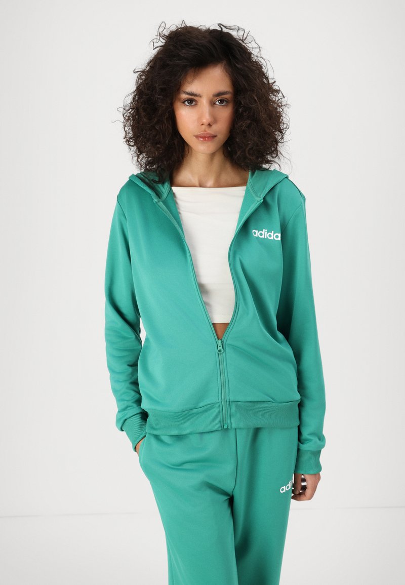 Felpa con zip color teal, dotata di cappuccio foderato e polsini a coste, caratterizzata da un logo Adidas bianco sul lato sinistro del petto. Abbinata a pantaloni coordinati.
