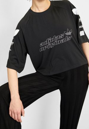 Osoba ubrana w czarny, oversize'owy t-shirt Adidas Originals z białym logo i pasujące czarne, teksturowane spodnie na jednolitym tle.