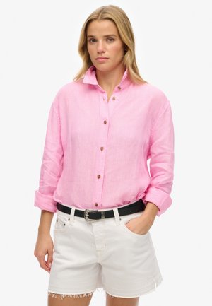 Chemise boutonnée en lin rose clair avec un col, des manches retroussées et des boutons marron, associée à un short en denim blanc et une ceinture noire.
