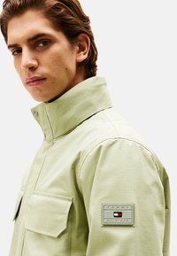 Chaqueta verde clara con un alto cuello, bolsillo en el pecho y tejido texturizado. Presenta un parche con el logo de Tommy Hilfiger en el brazo izquierdo.