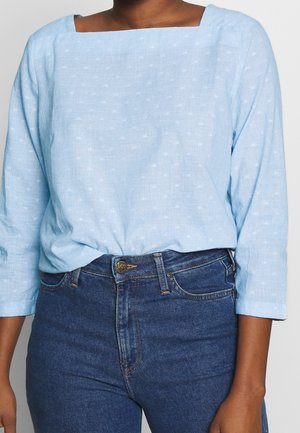 Blouse en coton bleu clair avec un décolleté carré, des manches longues et une texture subtile. Associée à un jean denim bleu foncé taille haute.