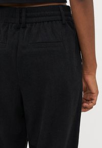 Black corduroy trousers with a textured finish, featuring an elastic waistband and rear pockets, shown from the waist down.