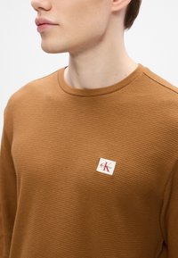 Καφέ πουκάμισο με φανέλα, μακρυμάνικο, με στρογγυλή λαιμόκοψη και υφή στην επιφάνεια. Διαθέτει μια μικρή, επώνυμη ραφή στην επάνω αριστερή γωνία.