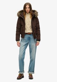 Bruine pufferjack met een met bont gevoerde capuchon, crèmekleurige gebreide trui, lichtblauwe losse jeans en bruine veterlaarzen met gele veters.