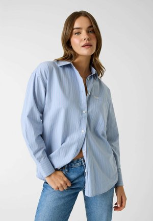 Giovane donna con capelli castano chiaro che indossa una camicia larga a righe azzurre e jeans blu su uno sfondo semplice.