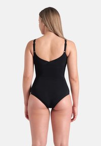Mulher vestindo um fato de banho preto de uma peça com alças ajustáveis, vista de costas contra um fundo claro e simples.