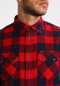 Camisa de franela a cuadros en rojo y azul marino con cuello abotonado, dos bolsillos frontales con solapas y botones oscuros en contraste.