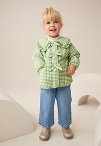 Hellgrüne Steppjacke mit kariertem Muster, Rüschenkragen, Schleifendetails und elastischen Bündchen, kombiniert mit weitem Jeanshose und grauen Stiefeln.