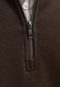 Metallglidelås delvis åpen på et brunt strikket plagg, som avslører en hvit skjorte med en synlig knapp under.